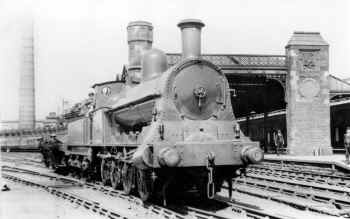 LNWR "A" class 0-8-0  engine number 1824  at south end of  Carnforth yard and station. The LNWR Coat of arms (Britannia) can be seen above the clock on the pillar supporting the station roof.  The Iron Works Chimney can also be seen clearly in the background. Cumbrian Railway Association Bowtell Collection.